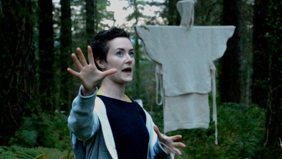 A lady, with short dark hair, standing in a forest, making hand gestures that indicate she’s discussing the scene around her, while in the background there is a hooded, white cloth, suspended from one of the trees, looking a little bit like a ghost